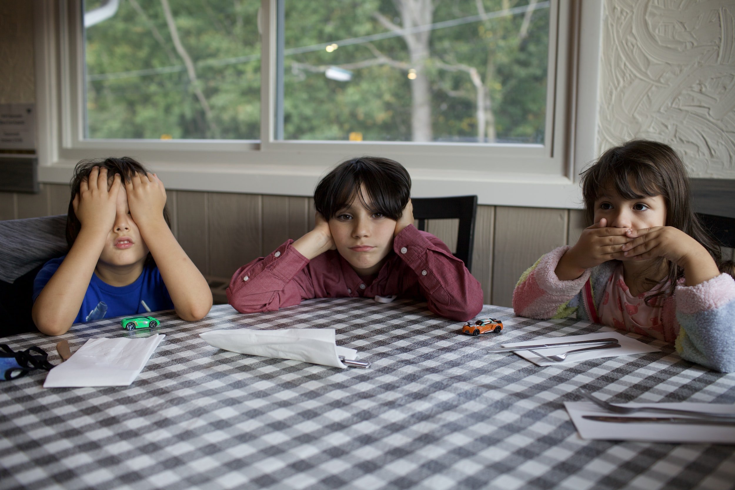 3 children sitting at table, one covering eyes, one covering ears, one covering mouth